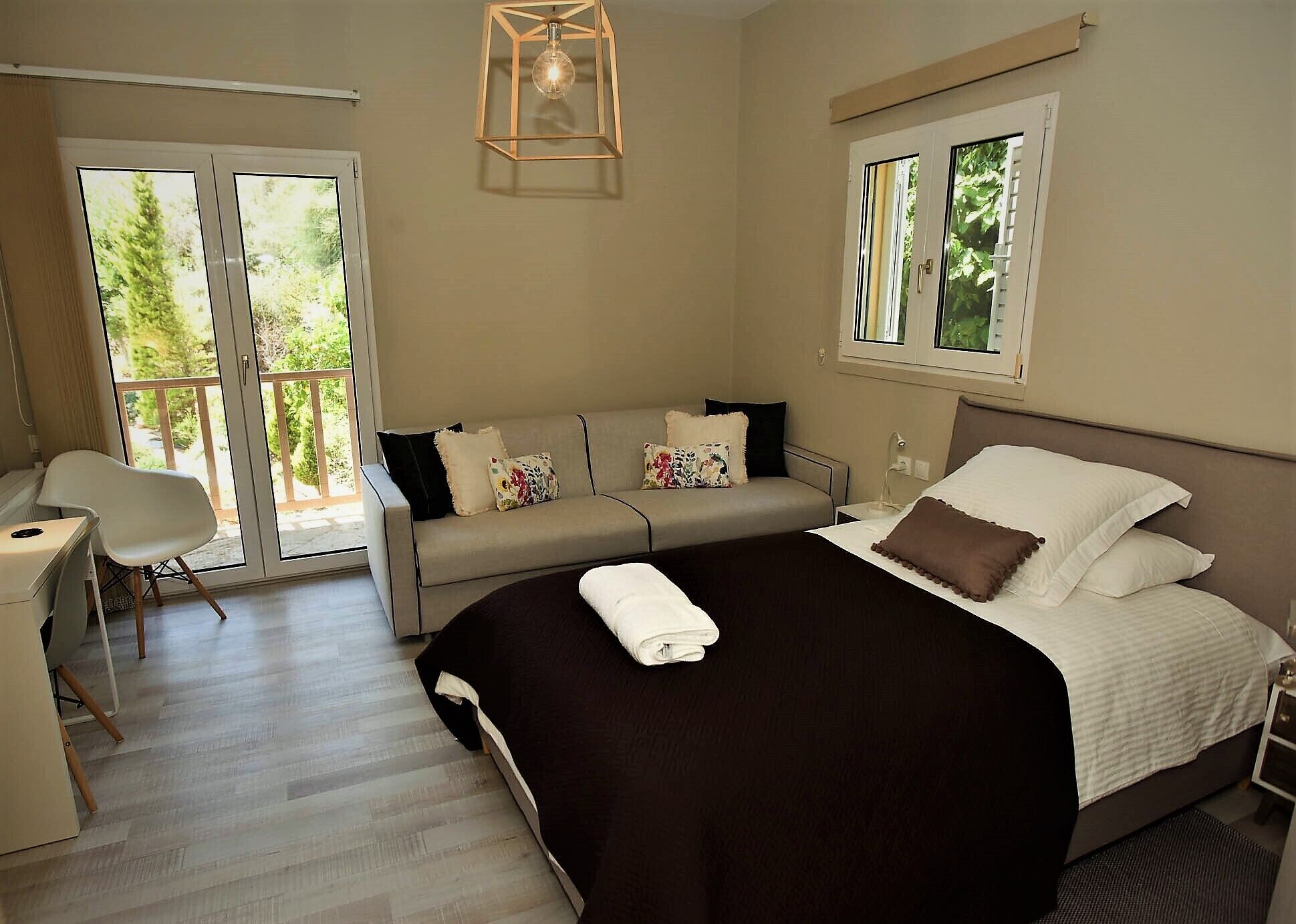 A bedroom with a bed, a sofa, a desk, and a chair. There is a window with a view of greenery.
