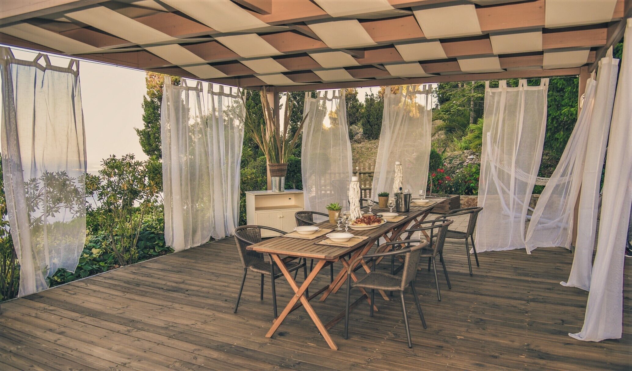 Un patio al aire libre con mesa de madera dispuesta para comer, rodeado de sillas y cubierto por una pérgola con cortinas blancas.