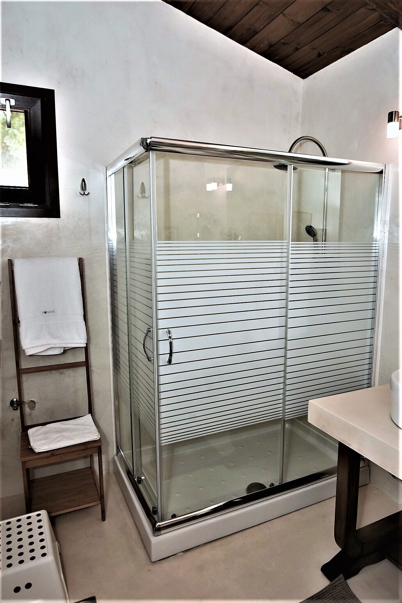 A glass-enclosed shower with a tiled floor, a wooden chair with a towel, and a white sink.