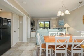 A compact kitchen with a refrigerator, a dining table with chairs, and a view of the outdoors through a window. - Tennis Villa E22 - 2BR Villa Steps to Beach (Hilton Head Island)