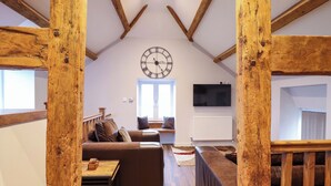 Ein gemütliches Loftsesselzimmer mit freiliegenden Holzbalken, einer großen Uhr, einem Sofa, einem kleinen Tisch und einem an der Wand befestigten Fernseher.