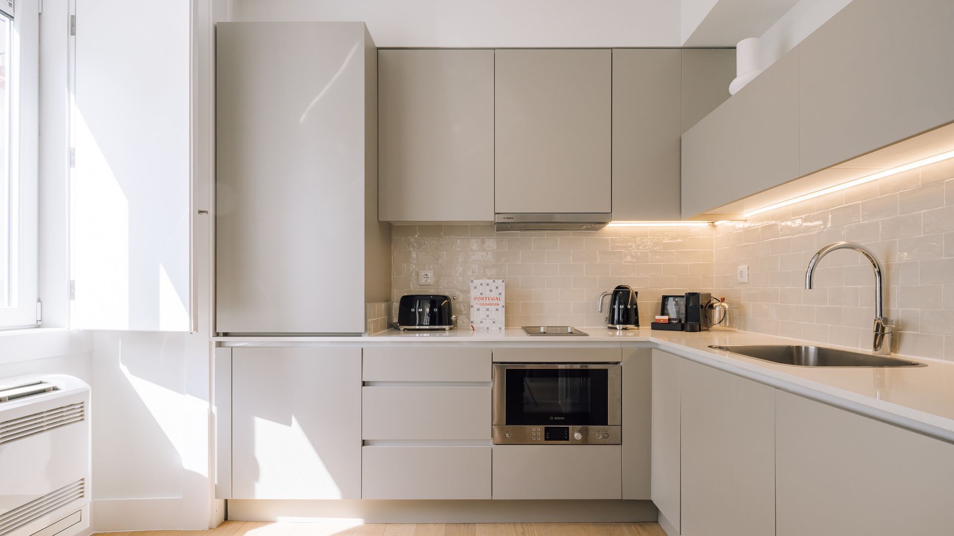 Une cuisine moderne avec des meubles blancs, un four Bosch, une machine à café et un livre de recettes portugaises posé sur le plan de travail.