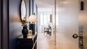 Ein Flur mit dunkler Wand, einem Spiegel, einer Vase mit getrockneten Blumen, einer Lampe und einer Tür zu einem Raum mit Tisch und Stühlen.