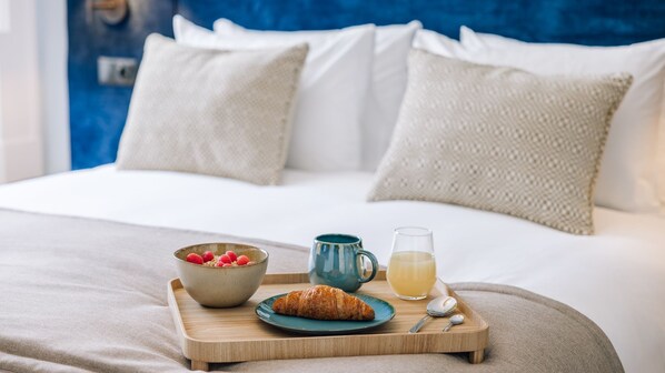 Ein ordentlich bezogenes Bett mit weißer Bettwäsche und zwei Kissen, ein Holztablett mit Frühstücksangeboten, darunter ein Croissant, eine Schale Himbeeren, eine Tasse und ein Glas Saft.