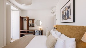 Habitación de hotel con cama, escritorio, silla, armario, espejo, lámpara y un cuadro en la pared.