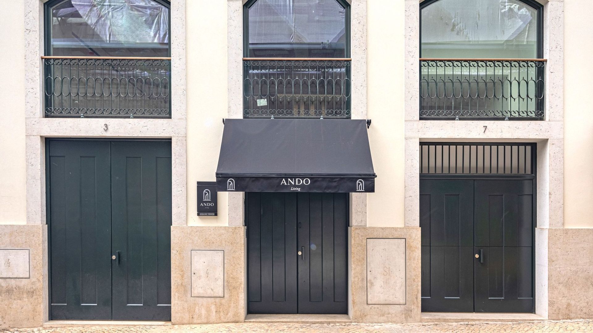 A building facade with three black double doors, each marked with a number (3, 4, 7). Above the central door, there is a black awning with the text "ANDO Living".