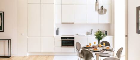 A modern kitchen with white cabinetry, a built-in oven, and a dining area with a round table and wire chairs.