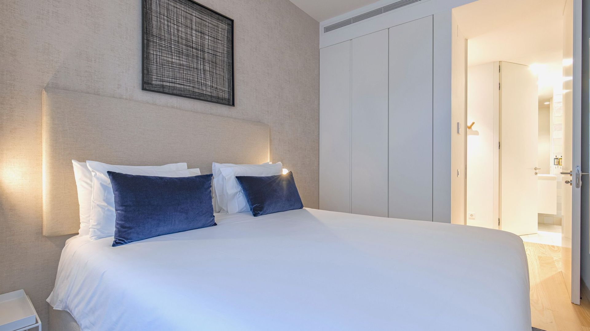A hotel room with a large bed, white linens, two dark blue pillows, a headboard, a wall-mounted artwork, and a wardrobe.