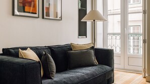 A dark grey sofa with multiple cushions, a floor lamp, and two framed artworks on the wall. - Ando Living - São Nicolau House - 101 (Lisboa)