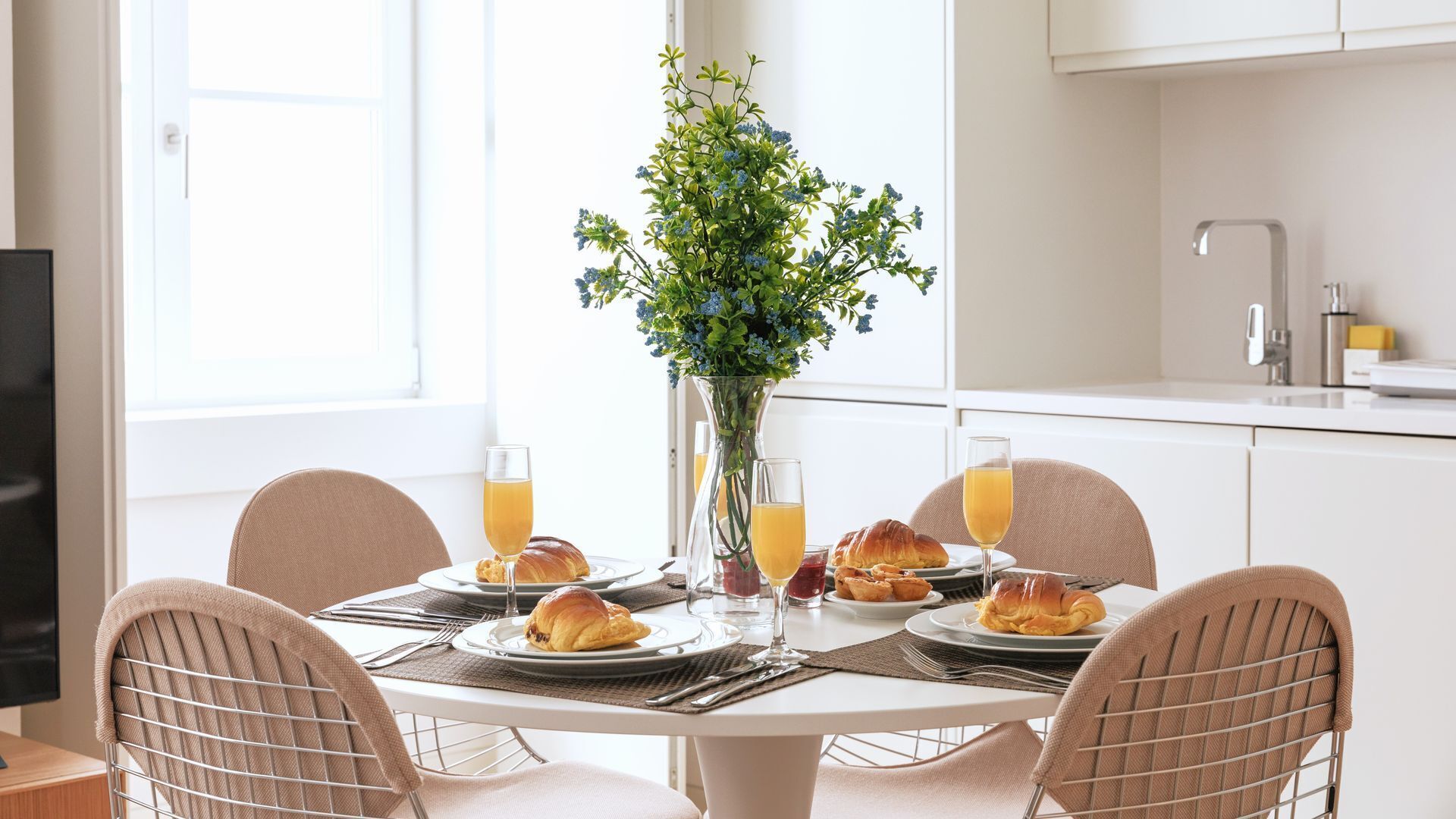 Un desayuno con una mesa, sillas, un jarrón con flores y platos con cruasanes y vasos de jugo.
