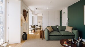 A modern living room with a green sofa, a dining area with a table and chairs, and a kitchen in the background. - Ando Living - One Bedroom Apartment with City View (Lisbon)