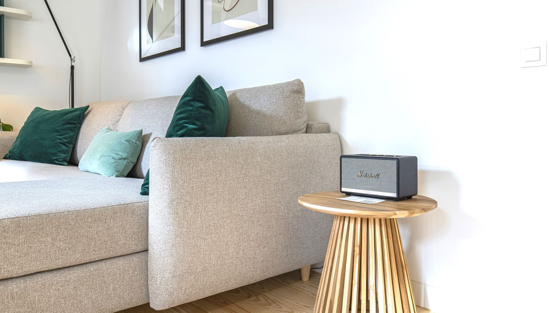 A modern living room with a grey sofa, green and teal cushions, a small wooden side table with a speaker, and a framed picture on the wall.