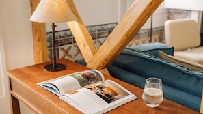 A wooden side table with a lamp, a book, a glass of water, and a blue sofa in a room with wooden beams and patterned wallpaper. - Ando Living - Santa Justa 79 House - 201 (Lisboa)