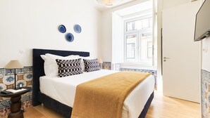 Quarto com cama, mesa de cabeceira, abajur e janela com vista para edifícios.