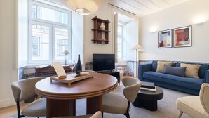 Uma sala de estar moderna com uma mesa de madeira redonda, duas cadeiras, um sofá azul, uma televisão e uma estante.
