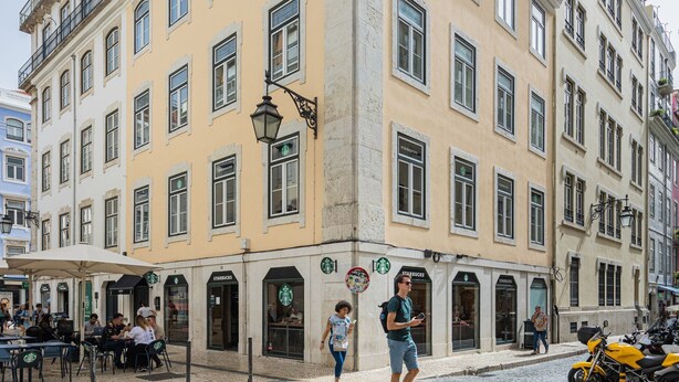 Um edifício em esquina com várias lojas Starbucks, mesas ao ar livre e rua de paralelepípedos.
