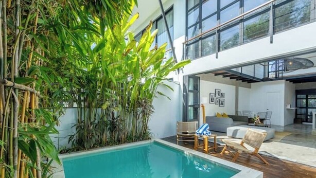 An indoor swimming pool with bamboo plants, a modern living area with a sofa and chairs, and large windows. - Urban Oasis Loft by GORO (Kabupaten Badung)