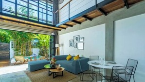 A modern living room with a blue sofa, a coffee table, and a dining area with a white table and black chairs. There are framed pictures on the wall and a view of greenery outside. - Urban Oasis Loft by GORO (Kabupaten Badung)
