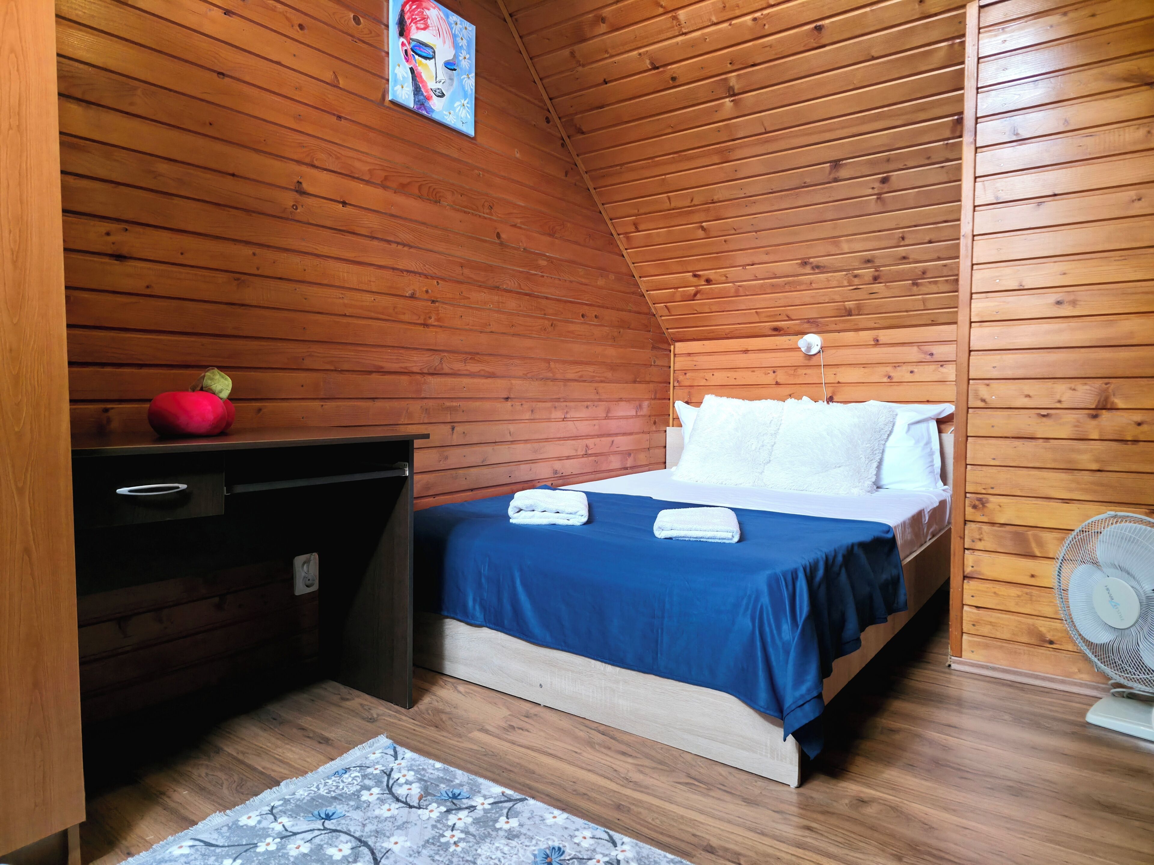 A bedroom with a wooden ceiling, a bed with white pillows, a blue bedspread, a desk with a red object on it, and a fan.