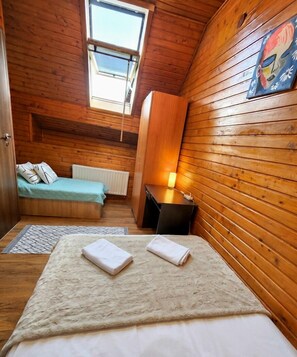 A cozy room with wooden walls, a bed with a white duvet, a small table with a lamp, a window, and a radiator.