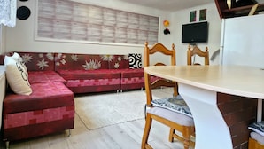 Una habitación con una mesa de comedor, un sofá de tapicería floral, un televisor montado en la pared y una persiana con estampado.