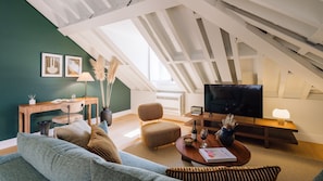 A cozy loft-style living room with a high ceiling, a flat-screen TV, a comfortable sofa, a wooden coffee table, and a dining table with chairs.