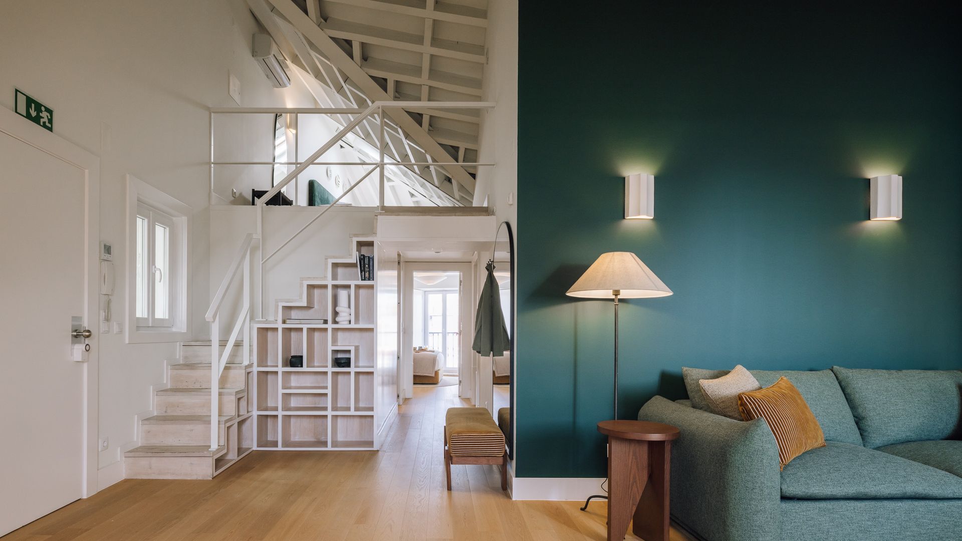 A staircase leading to an upper floor, a teal-colored wall with two wall-mounted lights, a green sofa with cushions, and a wooden bench.