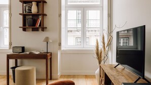 Ein Wohnzimmer mit einem Holzboden, einem Fernseher auf einem TV-Schrank, einem kleinen Tisch mit Lampe, einem Bücherregal und einem Fenster mit Blick auf Gebäude.