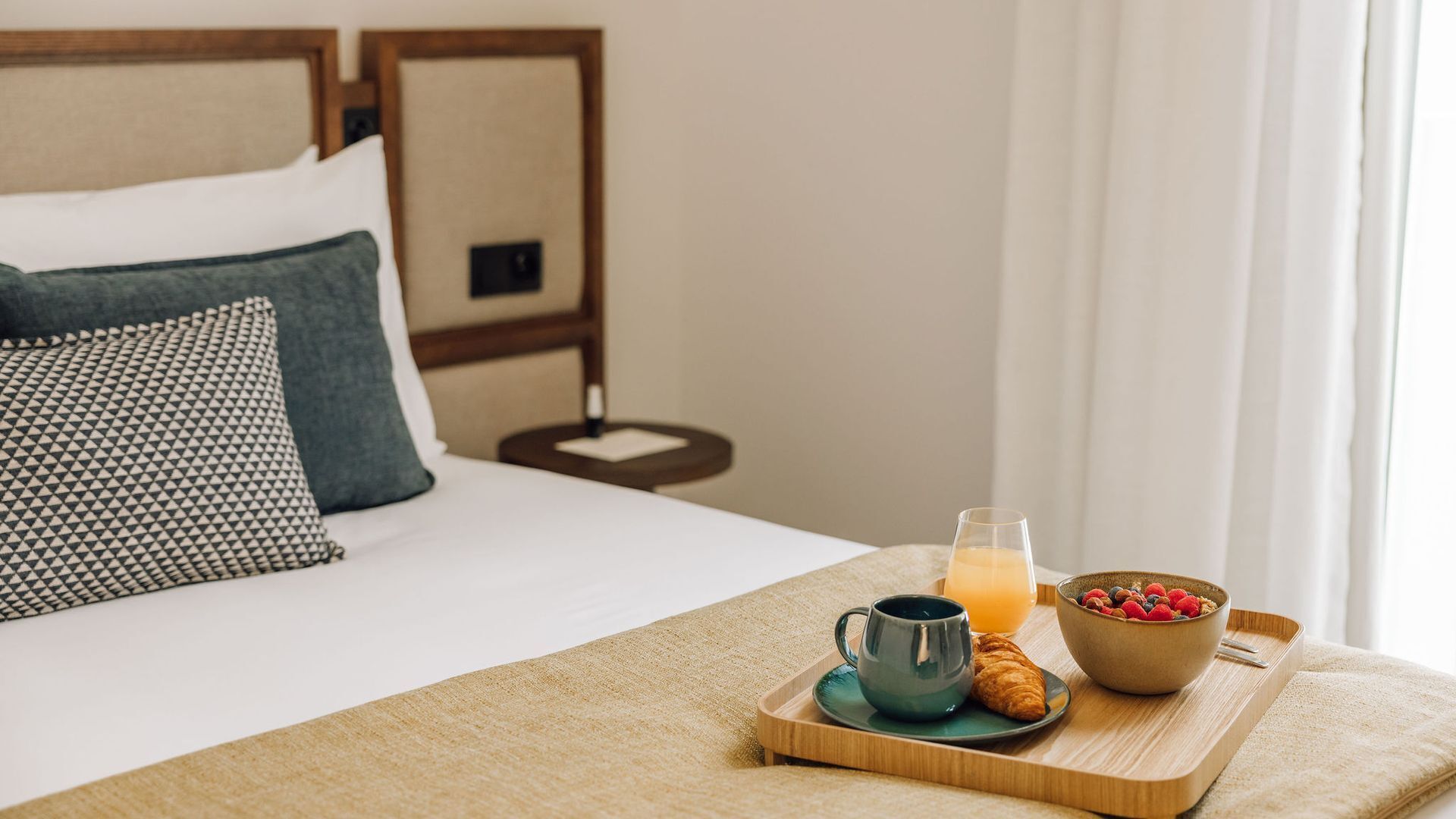 A neatly made bed with a wooden headboard, a bedside table with a lamp, and a breakfast tray with a croissant, mug, glass of orange juice, and a bowl of berries.