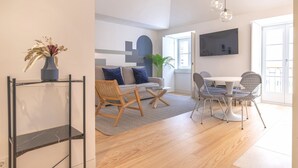 Ett modernt vardagsrum med en soffa, ett soffbord och en matplats med stolar. En tv är monterad på väggen och det står en växt i hörnet.
