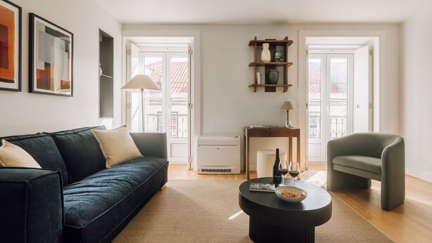Ett modernt vardagsrum med en svart soffa, en grå fåtölj och ett soffbord. Det finns ett litet sidobord i trä med en lampa och en flaska. Rummet har stora fönster med utsikt över ett tak täckt med röda tegelpannor.