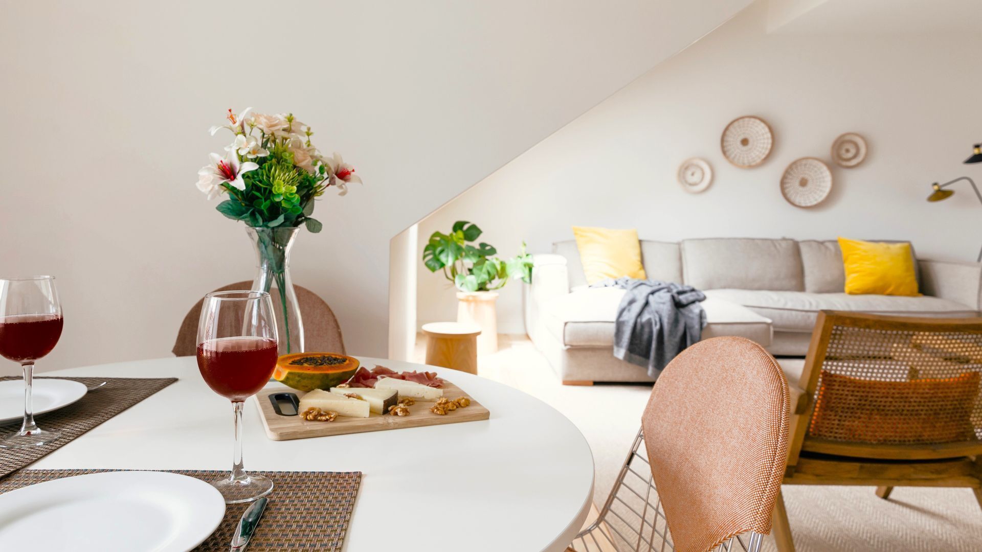 A dining area with a white table set for two, featuring a glass of red wine, a vase with flowers, and a wooden board with cheese and walnuts.