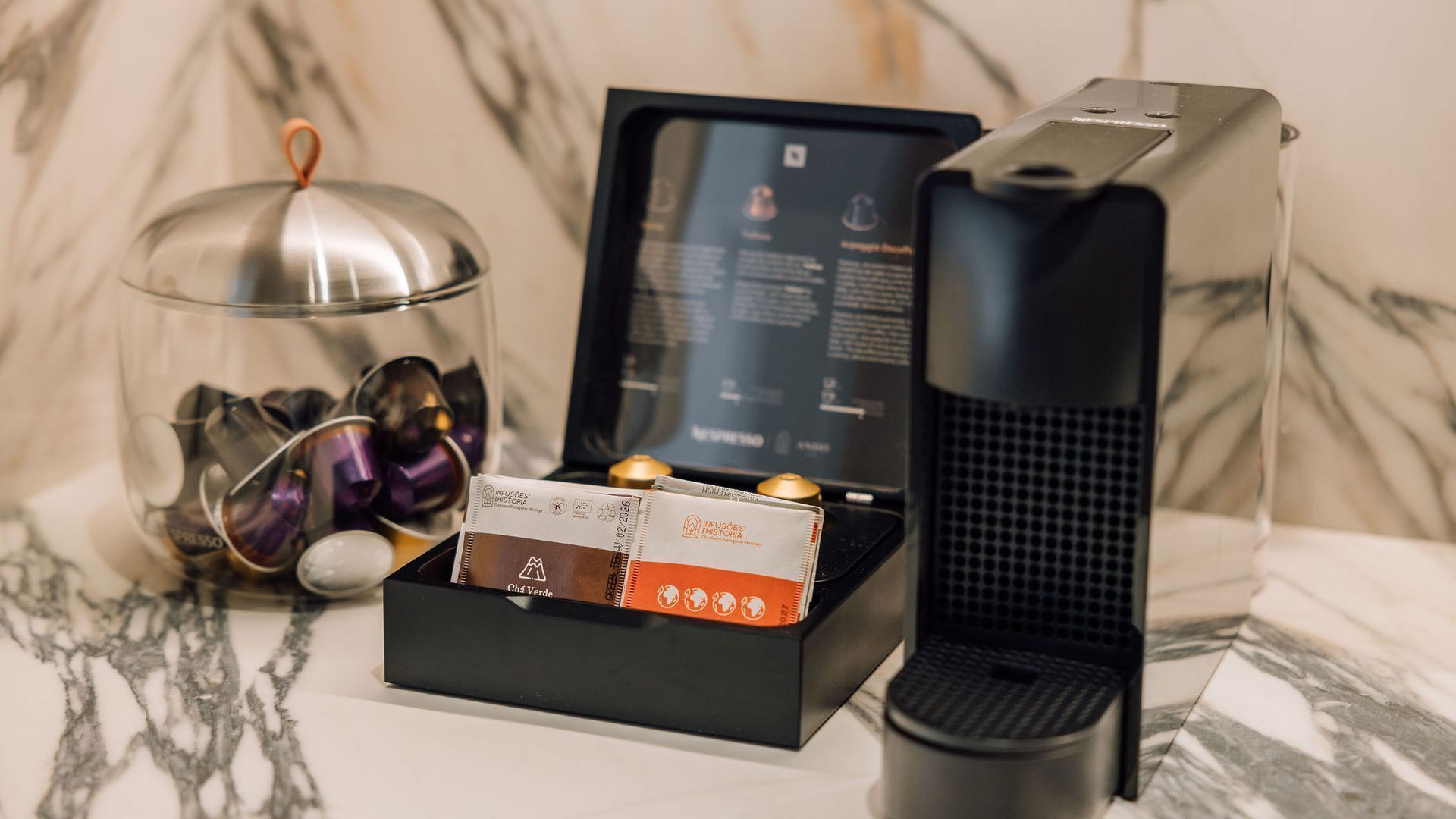 A coffee machine, a glass container with coffee capsules, and a box of coffee pods on a marble surface.