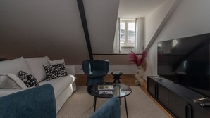 Een moderne woonkamer met een bank, een blauwe fauteuil, een glazen salontafel, een televisie en een raam met gordijnen.