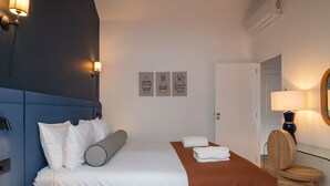 Hotelkamer met een blauw hoofdbord, witte beddengoed, een bruin plaid en een houten nachtkastje met een lamp.