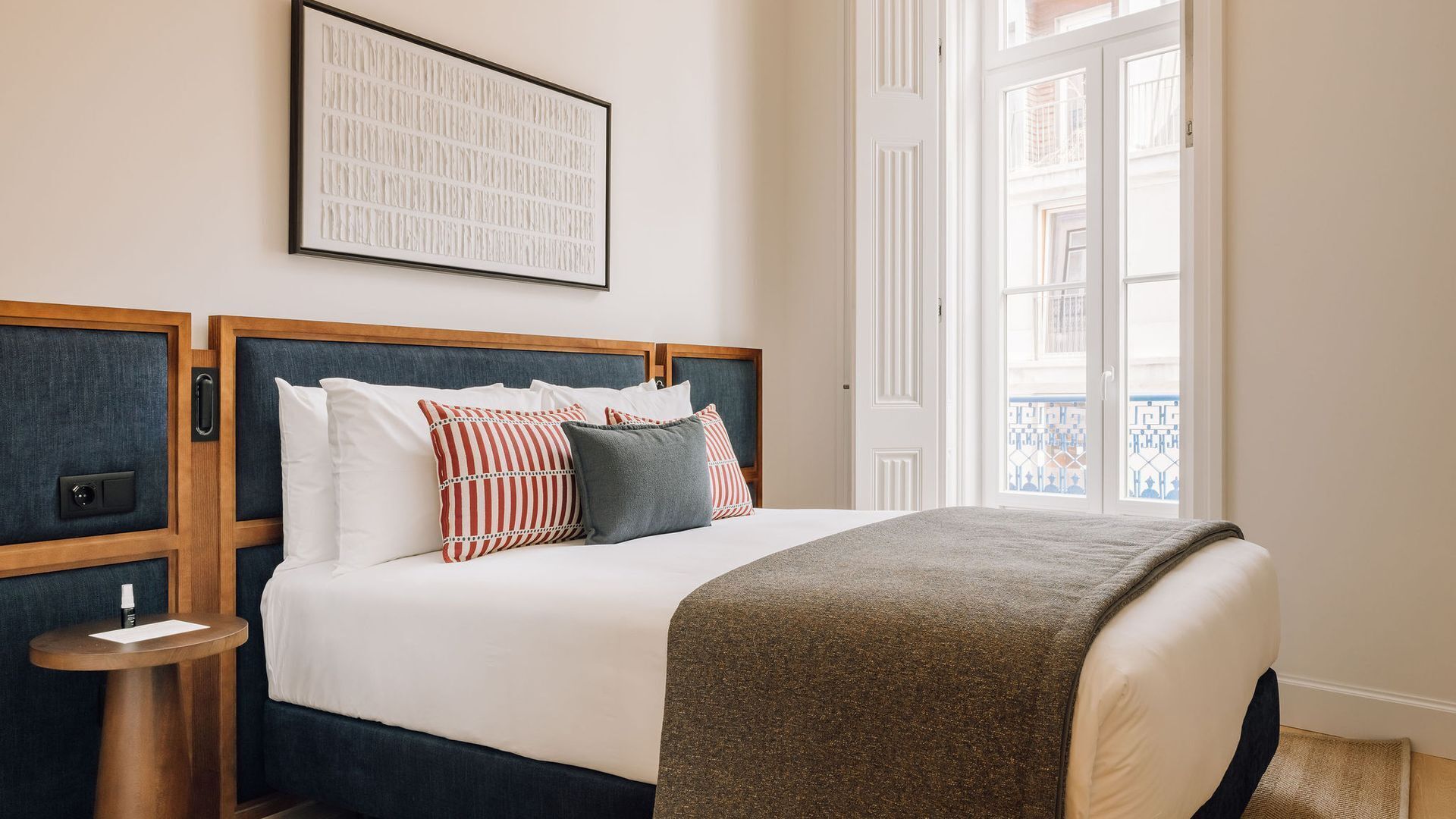 Una cama bien hecha con un cabecero oscuro, ropa de cama blanca y una variedad de almohadas. Hay una mesita de noche con una lámpara y un cuadro enmarcado sobre la cama.