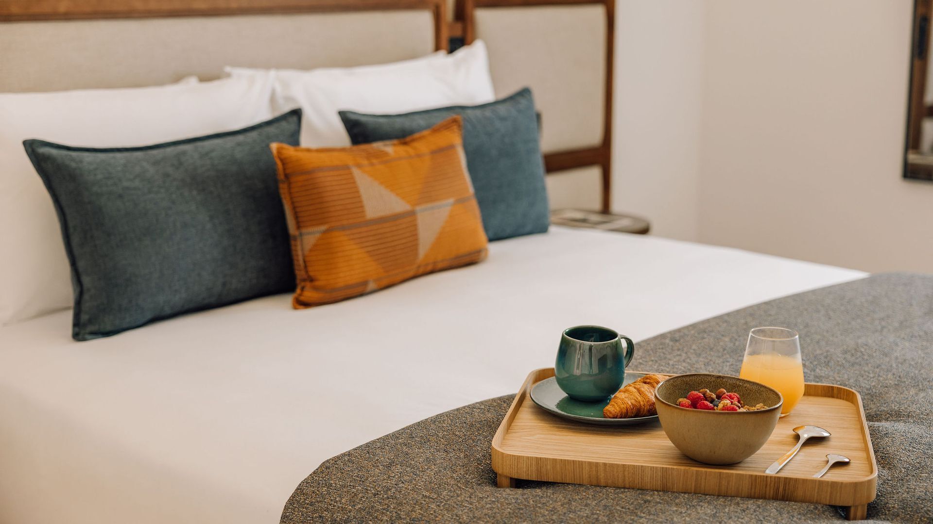 A neatly made bed with a wooden headboard, a tray with breakfast items including croissants, a bowl of berries, a mug, and a glass of orange juice.