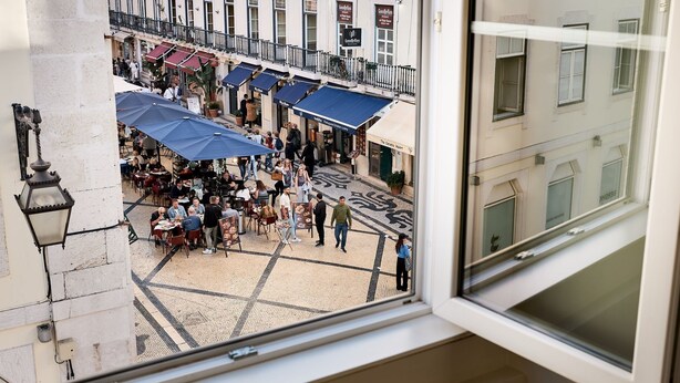 Um café ao ar livre com mesas e guarda-sóis, rodeado por edifícios com varandas e placas.