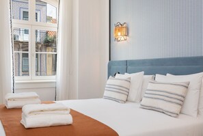 A neatly made bed with white linens and striped pillows, a blue headboard, and a window with curtains. - Ando Living - Santa Justa 77 House - 301 (Lisboa)