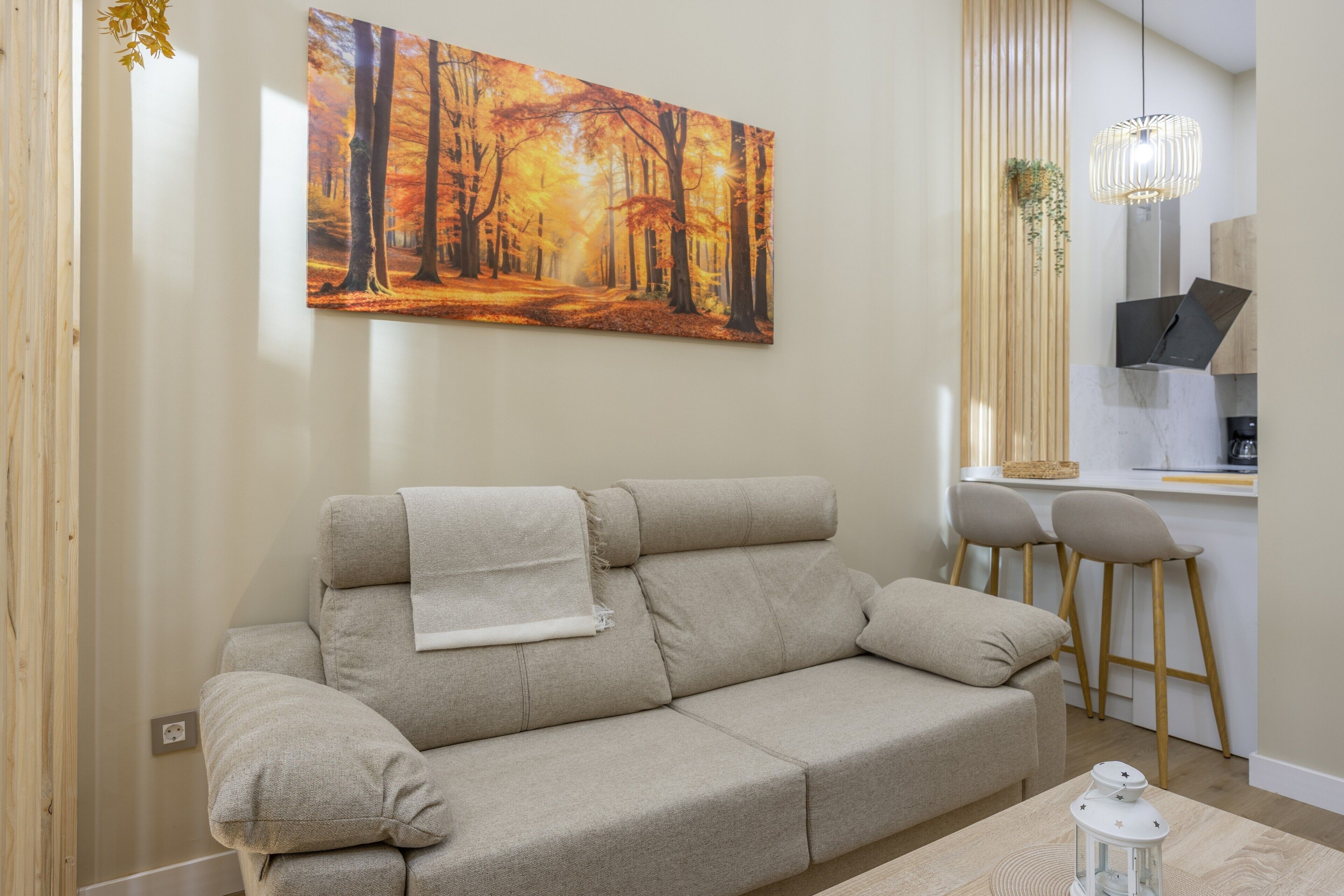 A living room with a sofa, a painting of a forest, a hanging light fixture, and a small table with a lantern.