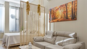 Une pièce avec un canapé beige, une table en bois sur laquelle est posée une lanterne, un lit recouvert d’une courtepointe blanche et un tableau représentant une forêt.