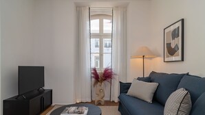 Un salon avec un canapé bleu foncé, une table basse noire sur laquelle repose un magazine, un téléviseur posé sur un meuble noir, un vase avec des fleurs rouges, une lampe et un tableau encadré accroché au mur.