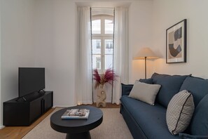 Ein Wohnzimmer mit einem dunkelblauen Sofa, einem schwarzen Couchtisch mit einer Zeitschrift, einem Fernseher auf einem schwarzen Lowboard, einer Vase mit roten Blumen, einer Lampe und einem gerahmten Bild an der Wand.