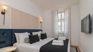 Une chambre d’hôtel avec un grand lit, une table de chevet, un téléviseur fixé au mur et une fenêtre avec des rideaux légers.