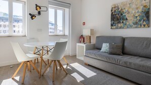 A modern living room with a grey sofa, white chairs, a round dining table, and a geometric painting on the wall. - Garcia 28 | Apartment 1B by LovelyStay (Lisboa)