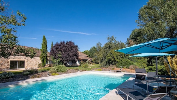 Ein Swimmingpool mit Liegestühlen und Sonnenschirmen, umgeben von Steinbauten und Grünflächen.