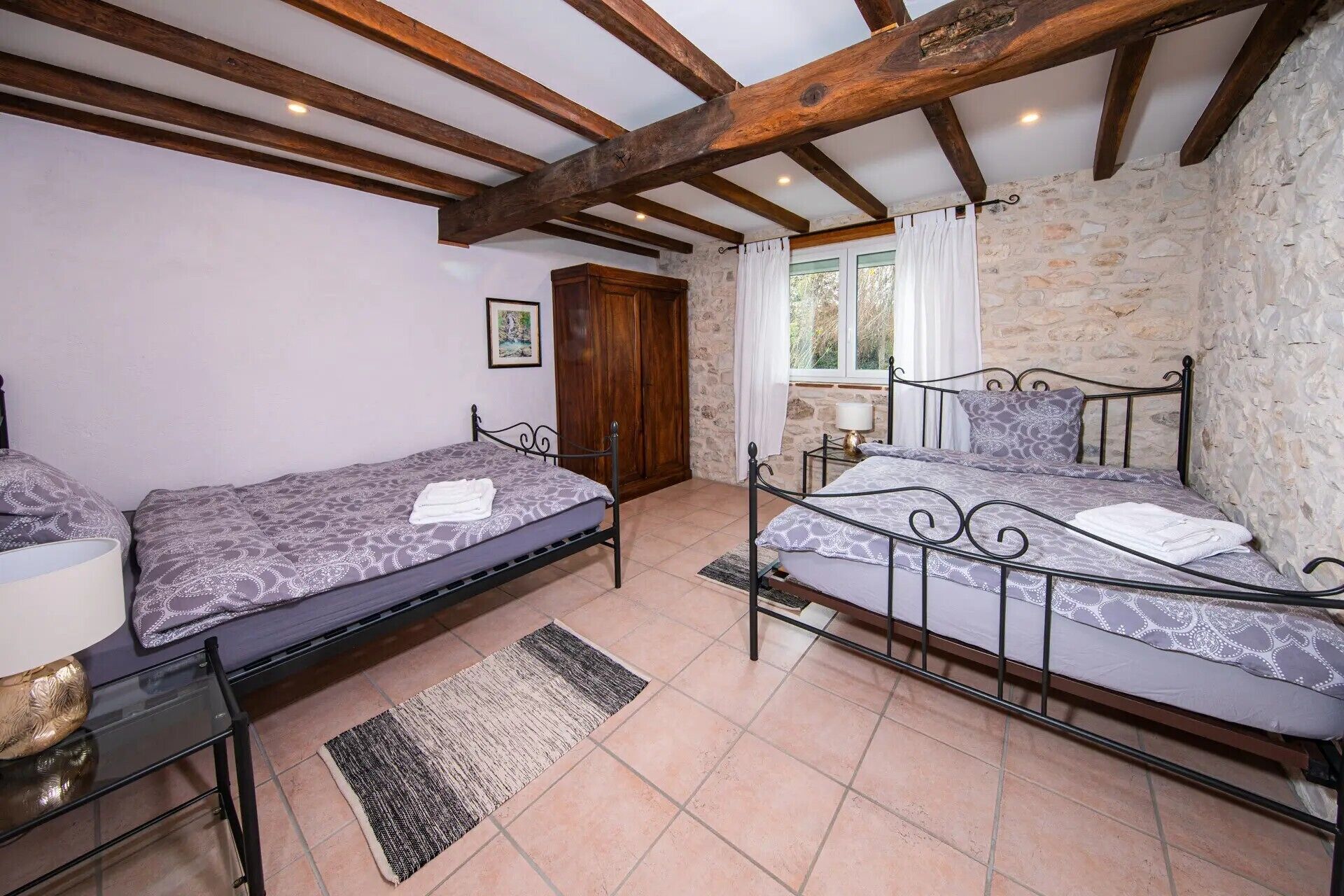 Une chambre avec deux lits, un mur en pierre, des poutres en bois, une fenêtre avec des rideaux et un tapis.