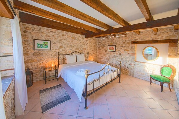 A bedroom with a stone wall, a bed with a metal frame, a nightstand, a green chair, and a window with a mirror.