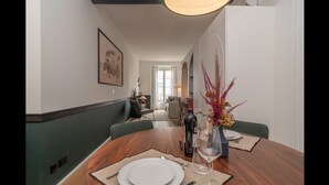 A dining area with a wooden table set for two, a vase with dried plants, and a bottle of wine. A hallway leads to a living area with a sofa and a lamp. - Ando Living - Santa Justa 77 House - 401 (Lisboa)