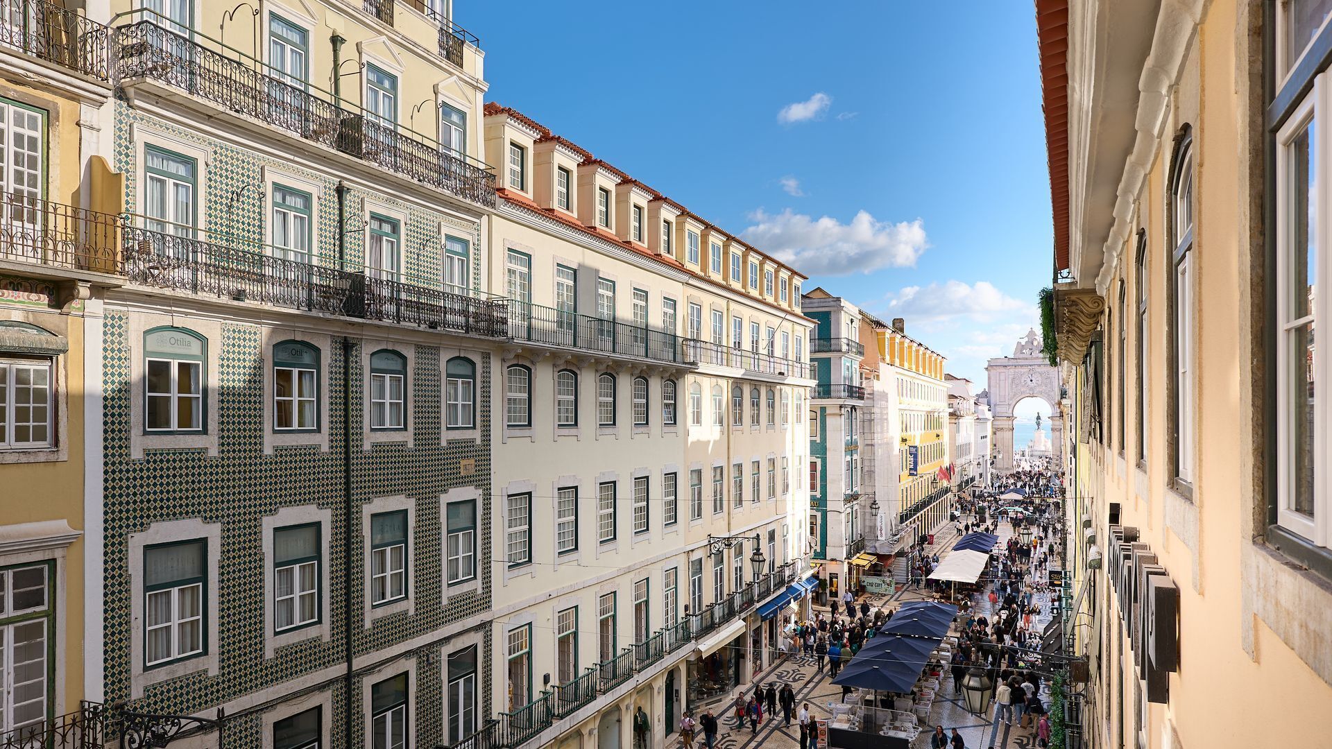 Uma rua movimentada com edifícios históricos, uma arcada imponente e áreas de estar ao ar livre.
